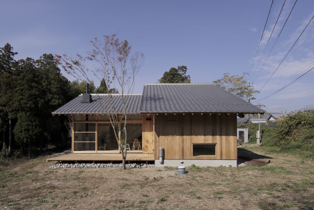 ザイツ建築の作品「てるおかの家」外観南側から北側を見る写真、南面に３枚の大きなガラスがありその前には広いウッドデッキそのウッドデッキの前には家の高さを超えるほどの高い楓の植林。外壁は大分県日田市産の日田杉の赤身のみ使用した加工板を縦に使用、同じ材で開き窓も１箇所、瓦屋根はいぶし銀で薪ストーブの煙突が伸びている。