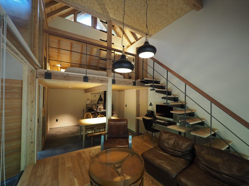 ザイツ建築が手がけた「蓑帽子の家」の吹き抜けのあるリビングより二階への階段、手すりはアイアン製作で手元はブビンガ材を使用、2階は寝室でかすかにベッドが見える。リビング横のダイニングは玄関ドアがありそのまま土足のまま使用ダイニングにはドミノ8クッキングストーブがある